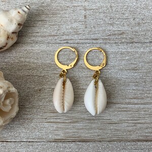May include: A pair of gold-colored hoop earrings with a natural seashell pendant. The earrings are displayed on a weathered, gray wooden surface, with seashells in the background. The earrings have a beachy, coastal theme.