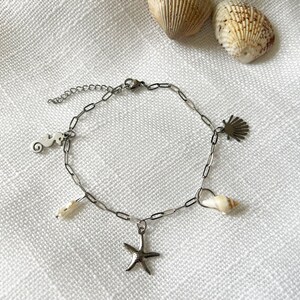 Peut inclure: Un bracelet à breloques argenté avec des breloques en forme d'hippocampe, d'étoile de mer et de coquillage. Le bracelet est présenté sur une surface blanche texturée avec des coquillages en arrière-plan. Un bijou délicat.