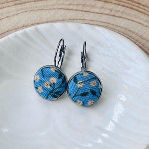 Puede incluir: Un par de pendientes colgantes redondos con una incrustación de tela azul brillante con un patrón de pequeñas flores blancas y hojas de color verde oscuro. Los pendientes están montados en un marco de metal oscuro con cierres de palanca.