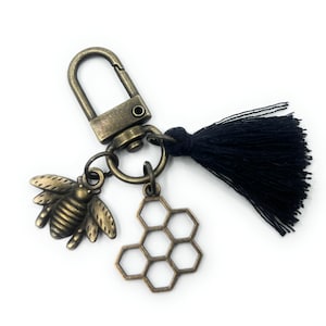 May include: A bronze-coloured keychain with a bee and honeycomb charm, a black tassel, and a clasp. The bee charm has detailed wings and body segments. The honeycomb charm has a hexagonal design. The keychain is on a white background.