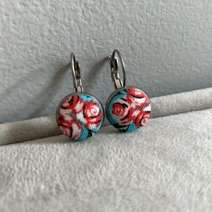May include: A pair of round leverback earrings. Each earring has a fabric-covered button with a pattern of red swirls on a light blue background, set in a silver-tone metal frame. The earrings are displayed on a light grey surface.