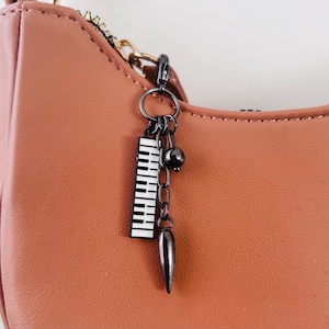 Puede incluir: Un bolso de mano color rosa empolvado con un dije de metal oscuro en la cremallera. El dije consta de un colgante rectangular en blanco y negro con teclas de piano, una pequeña esfera de metal oscuro y una punta metálica.