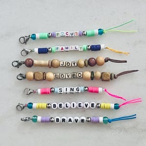 May include: Seven beaded zip pulls featuring silver lobster clasps and colourful disc or wooden beads. Each pull displays a white cube bead sequence spelling the words FOCUS, FAMILY, JOY, LOVED, SING, BELIEVE, and BRAVE with attached coloured cords.