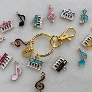 Puede incluir: Un llavero de metal dorado con cierre de mosquetón, que incluye un pequeño dije circular con la letra O. Alrededor del anillo hay varios dijes de esmalte con formas de notas musicales y teclados de piano en colores rosa, azul, negro y blanco.