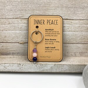 Puede incluir: Un llavero de metal con tres cuentas en morado, rosa claro y azul oscuro, sujeto a una tarjeta de presentaci&oacute;n con el t&iacute;tulo INNER PEACE. La tarjeta enumera amatista, cuarzo rosa y lapisl&aacute;zuli con sus propiedades y el nombre de la marca.