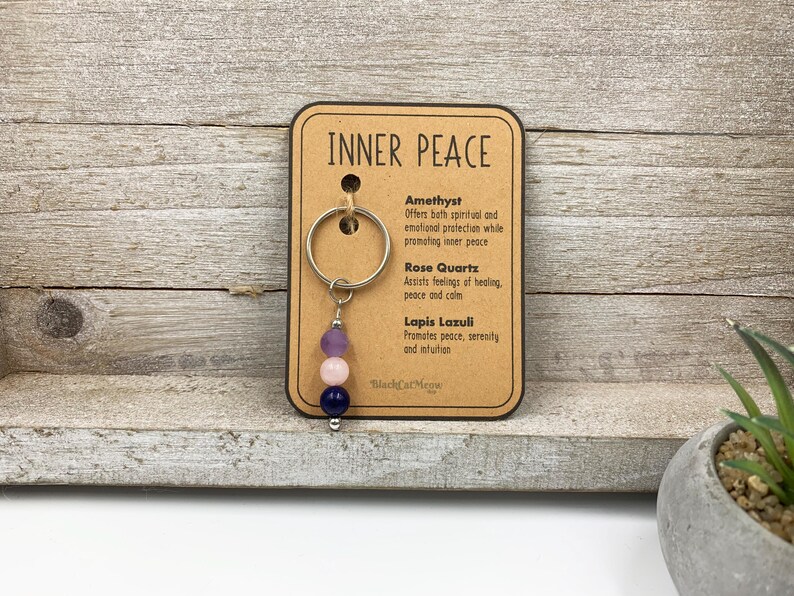 Puede incluir: Un llavero de metal con tres cuentas en morado, rosa claro y azul oscuro, sujeto a una tarjeta de presentaci&oacute;n con el t&iacute;tulo INNER PEACE. La tarjeta enumera amatista, cuarzo rosa y lapisl&aacute;zuli con sus propiedades y el nombre de la marca.