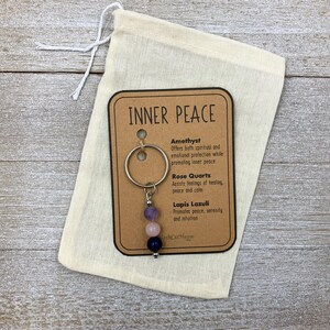 Puede incluir: Un llavero de metal con tres cuentas redondas de piedras preciosas en morado, rosa claro y azul oscuro, sujeto a una tarjeta con el texto INNER PEACE. La tarjeta enumera amatista, cuarzo rosa y lapisl&aacute;zuli, dentro de una bolsa de tela beige.