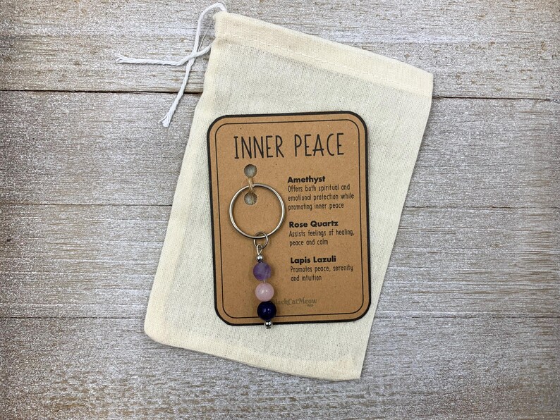 Puede incluir: Un llavero de metal con tres cuentas redondas de piedras preciosas en morado, rosa claro y azul oscuro, sujeto a una tarjeta con el texto INNER PEACE. La tarjeta enumera amatista, cuarzo rosa y lapisl&aacute;zuli, dentro de una bolsa de tela beige.
