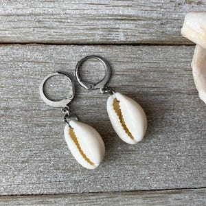 May include: A pair of earrings with natural white cowrie shells hanging from silver-coloured leverback closures. The earrings are shown on a weathered, grey wooden surface, with a seashell fragment in the background.