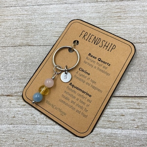 May include: A keychain with a charm featuring three gemstone beads in pink, yellow, and blue. The charm is attached to a silver key ring with a letter 'S' charm. A card with the text 'FRIENDSHIP' and descriptions of the gemstones Rose Quartz, Citrine, and Aquamarine is attached to the keychain.