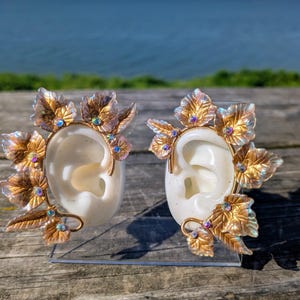 May include: A pair of ear-shaped earrings with gold-colored leaf accents and iridescent gems. The earrings are set against a blurred background of a body of water and a wooden surface.