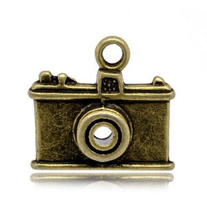 May include: A vintage-style camera charm made of metal with an antique gold finish. The charm has a detailed design with a lens, shutter, and a small loop at the top for attaching to a necklace or bracelet.