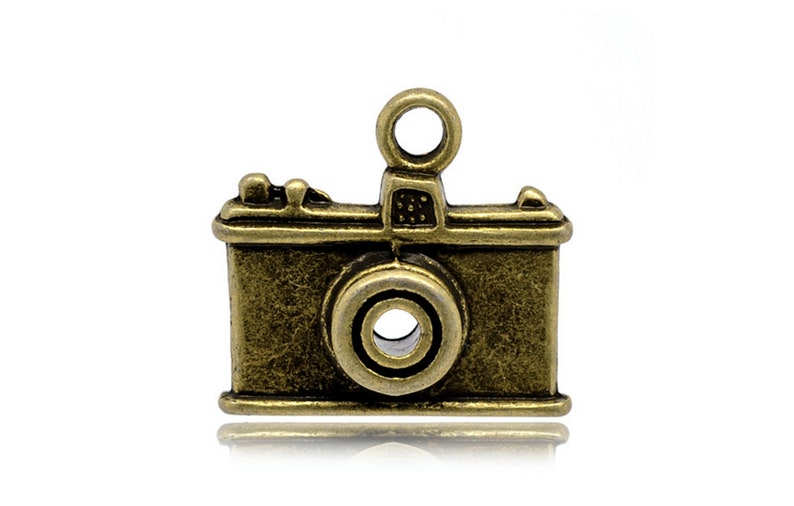 May include: A vintage-style camera charm made of metal with an antique gold finish. The charm has a detailed design with a lens, shutter, and a small loop at the top for attaching to a necklace or bracelet.