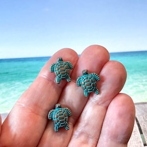 May include: Three small turquoise sea turtle charms with a bronze finish. The charms are on a hand against a blue ocean background.