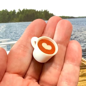 May include: A miniature white coffee cup with a handle, filled with a brown liquid and a white heart design. The cup is held in a person's hand, with a lake and trees in the background.