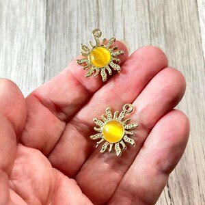 May include: Two gold-colored sun charms with yellow centers. The sunburst design features detailed rays around a central, round, yellow stone. Each charm has a small loop at the top for attaching to jewelry or other crafts.