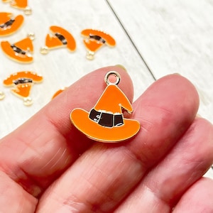 May include: A close-up of a gold-toned witch hat charm with an orange and black enamel design. The charm is being held in a hand.