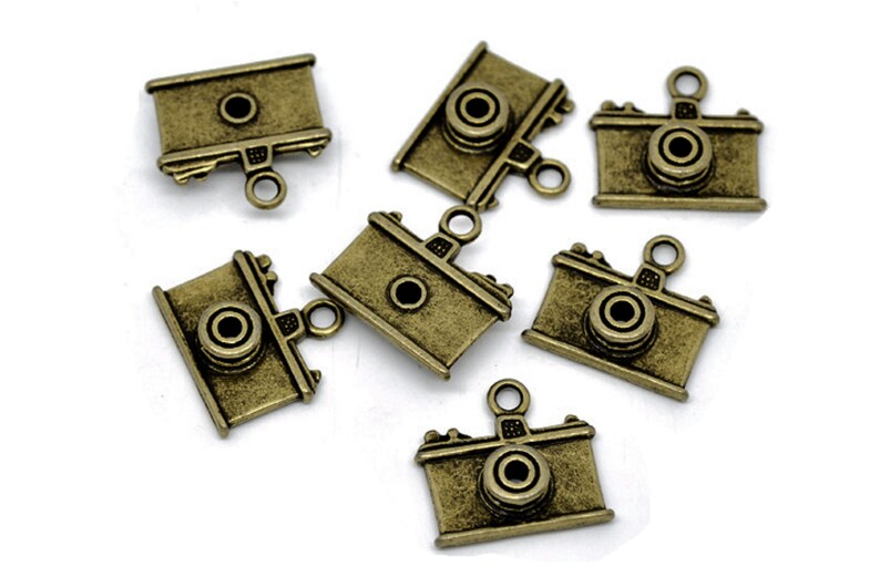 May include: Seven antique gold-toned camera charms with a loop for hanging. The charms are detailed with a lens and other camera features.