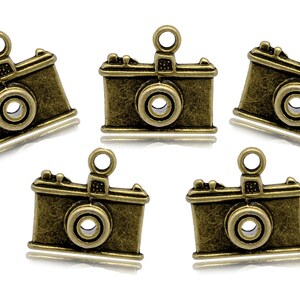 May include: Five antique gold-toned camera charms with a loop for hanging. The charms are detailed with a lens, a flash, and a textured surface.
