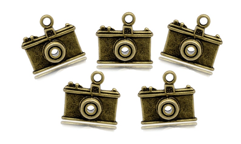 May include: Five antique gold-toned camera charms with a loop for hanging. The charms are detailed with a lens, a flash, and a textured surface.