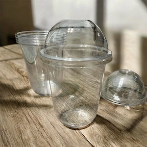 Könnte beinhalten: Klare Plastikbecher mit gewölbten Deckeln. Die Becher sind transparent und ermöglichen die Sichtbarkeit des Inhalts. Ein Becher hat einen Deckel, ein anderer einen Deckel in der Nähe, und ein dritter Becher ist im Hintergrund sichtbar.