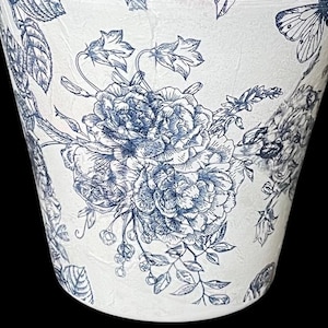 May include: A tapered cylindrical planter featuring a white base with a detailed navy blue botanical print. The design displays intricate illustrations of blooming peonies, delicate bell-shaped flowers, leafy stems, and a butterfly in flight.