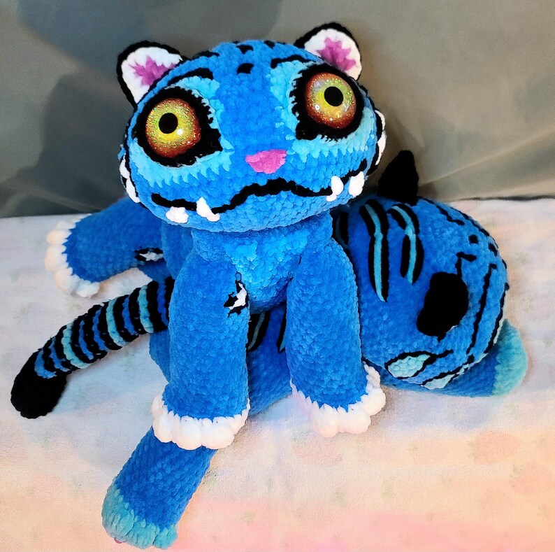 K&ouml;nnte beinhalten: Ein Pl&uuml;schtiger aus leuchtend blauem Garn mit schwarzen Streifen, gro&szlig;en gelben Glitzeraugen, einer kleinen rosa Nase und wei&szlig;en Akzenten an Ohren und Pfoten. Der Tiger hat ein breites, gesticktes Maul mit sichtbaren wei&szlig;en Z&auml;hnen und einen langen gestreiften Schwanz.