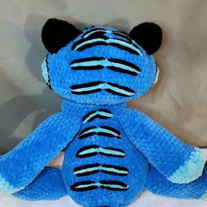 K&ouml;nnte beinhalten: Ein Pl&uuml;schtiger aus leuchtend blauem, strukturiertem Garn mit markanten schwarzen horizontalen Streifen auf Kopf, R&uuml;cken und Gliedma&szlig;en. Das Stofftier hat abgerundete schwarze Ohren sowie hellblaue Akzente an den Pfoten und der Schwanzspitze.