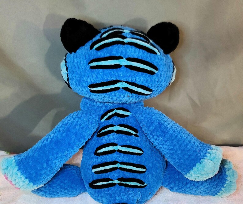 K&ouml;nnte beinhalten: Ein Pl&uuml;schtiger aus leuchtend blauem, strukturiertem Garn mit markanten schwarzen horizontalen Streifen auf Kopf, R&uuml;cken und Gliedma&szlig;en. Das Stofftier hat abgerundete schwarze Ohren sowie hellblaue Akzente an den Pfoten und der Schwanzspitze.