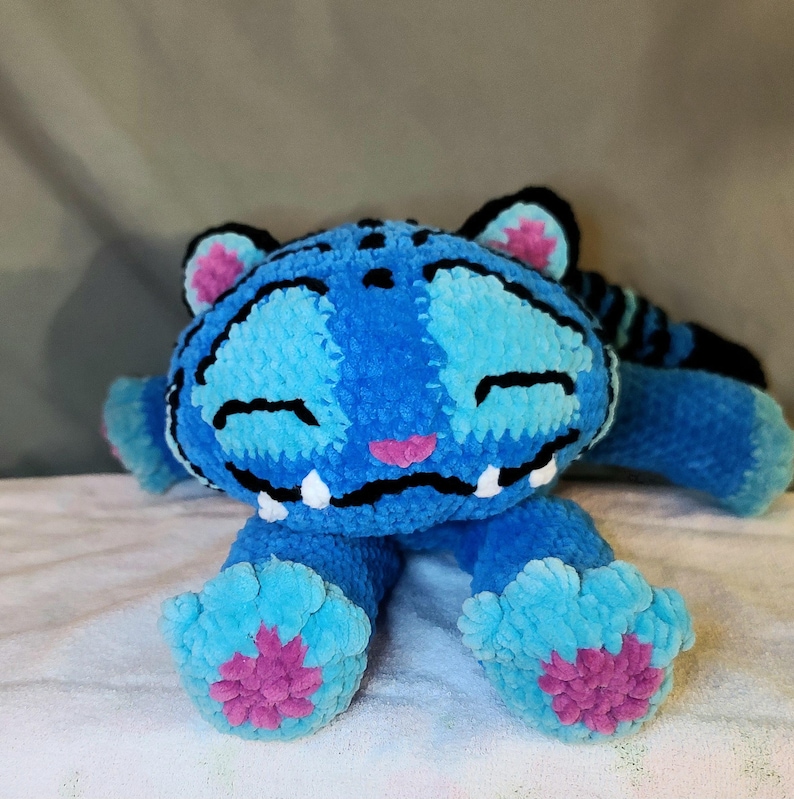K&ouml;nnte beinhalten: Ein geh&auml;keltes Tiger-Stofftier in Blaut&ouml;nen mit schwarzen Streifen, einer rosa Nase und rosa Pfotenballen. Die Figur hat ein breites, l&auml;chelndes Maul mit sichtbaren wei&szlig;en Z&auml;hnen, geschlossene Augen und abgerundete Ohren mit rosa Details.