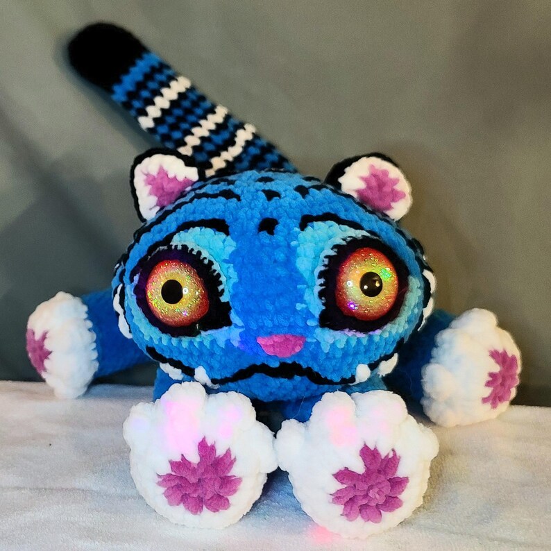 K&ouml;nnte beinhalten: Ein Pl&uuml;schtier in Form eines Wesens mit blau-schwarz gestreiftem Fell, gro&szlig;en irisierenden orangefarbenen Augen und einer kleinen rosa Nase. Es hat wei&szlig;e Pfoten mit rosa Akzenten, wei&szlig;e Innenohren und einen langen, gestreiften Schwanz.