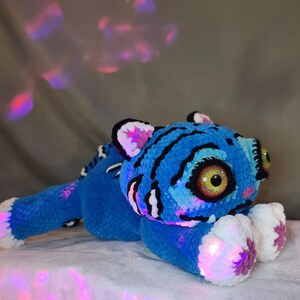 K&ouml;nnte beinhalten: Ein Pl&uuml;schtiger aus leuchtend blauem Garn mit schwarzen Streifen. Das Tier hat gro&szlig;e, runde, glitzernde goldene Augen, wei&szlig;e Pfoten mit rosa Akzenten und wei&szlig;e Innenohren. Das weiche, strukturierte Material reflektiert farbiges Licht.