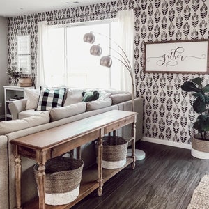 May include: A living room with a white sofa, a wooden console table with two woven baskets, and a black and white floral wallpaper with the word "gather" on a wooden sign.