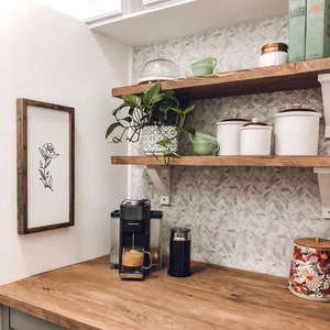 May include: A kitchen countertop with a coffee machine, a glass of coffee, and a canister with a floral design. The countertop is made of wood and has a light brown finish. The coffee machine is black and has a silver handle. The glass of coffee is clear and has a brown liquid inside. The canister is white with a floral design in red, orange, and yellow. The countertop is in front of a white tiled wall with a shelf above it. The shelf is made of wood and has a light brown finish. There are several items on the shelf, including a plant, a glass jar, and a white ceramic canister.