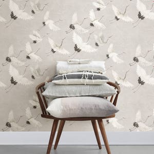 May include: A stack of pillows in various shades of gray and white, with a striped pattern, sits on a wooden chair against a wall with a wallpaper featuring a pattern of white cranes flying in different directions on a light beige background.