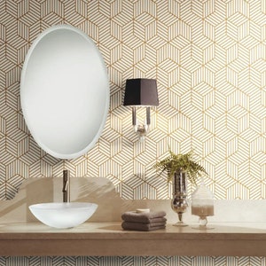 May include: A white bathroom with a gold geometric patterned wallpaper. The bathroom features a white oval mirror, a white porcelain sink, a silver faucet, a black lamp with a white shade, a brown towel, a clear glass jar, and a green plant in a silver vase.
