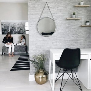 May include: A white desk with a black chair and a round silver mirror hanging on a grey wall. A vase with a gold finish holds a bouquet of white flowers.