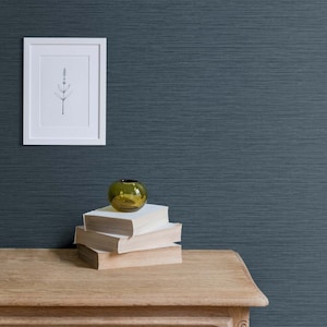 May include: A stack of three books with a small green glass vase on top, sitting on a wooden table in front of a dark blue textured wall. A framed black and white drawing of a plant is hanging on the wall.