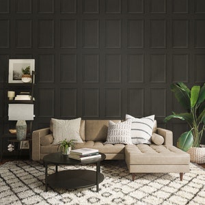Op de afbeelding: Een hoekbank met een lichtbruine lederen bekleding en een donkerhouten frame. De bank staat voor een muur met donkergrijs behang met een paneeldesign. De bank heeft twee kussens met een wit en grijs geometrisch patroon en een wit en zwart gestreept kussen. Een kleine zwarte salontafel met twee niveaus staat voor de bank. Een potplant staat op de salontafel. Een kleine potplant staat op een plank achter de bank. Een tapijt met een wit en grijs ruitpatroon bedekt de vloer.