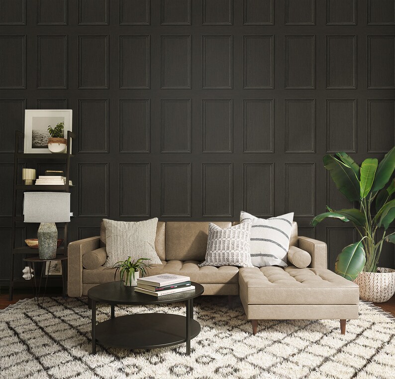 Op de afbeelding: Een hoekbank met een lichtbruine lederen bekleding en een donkerhouten frame. De bank staat voor een muur met donkergrijs behang met een paneeldesign. De bank heeft twee kussens met een wit en grijs geometrisch patroon en een wit en zwart gestreept kussen. Een kleine zwarte salontafel met twee niveaus staat voor de bank. Een potplant staat op de salontafel. Een kleine potplant staat op een plank achter de bank. Een tapijt met een wit en grijs ruitpatroon bedekt de vloer.