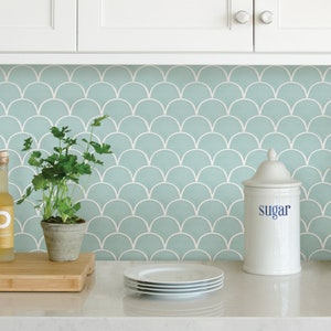 May include: A kitchen backsplash with a light blue and white scallop tile pattern. A white ceramic sugar jar with the word "sugar" printed on it is on the counter in front of the backsplash. There are also a few white plates and a potted plant on the counter.