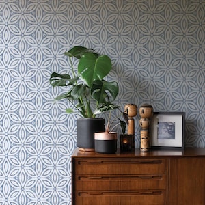 May include: A wooden chest of drawers displays potted plants, wooden dolls, a framed photo, and a candle. The backdrop is a blue and white floral wallpaper. The plants have large green leaves.
