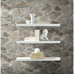 May include: Three white shelves against a backdrop of a stone wall wallpaper. The top shelf has a white vase and a small glass jar. The middle shelf has a white ceramic lion figurine and several books. The bottom shelf has a stack of books and a white ball.