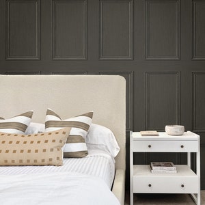 Op de afbeelding: Een wit bed met een wit hoofdeinde en een wit beddengoed. Er liggen drie kussens op het bed, twee gestreepte kussens en een effen kussen. Een wit nachtkastje met twee laden en een plank staat naast het bed. Op de bovenste plank van het nachtkastje staan een kleine kom en een boek. De muur achter het bed is bedekt met donkergrijze behang met een houten paneel ontwerp.