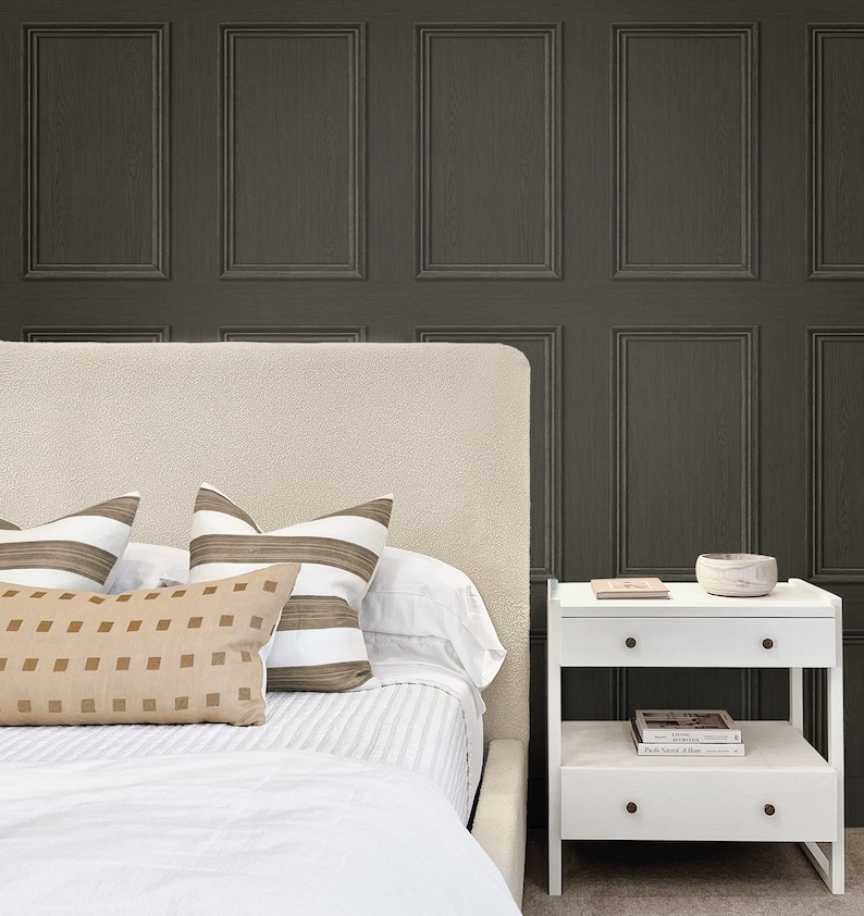 Op de afbeelding: Een wit bed met een wit hoofdeinde en een wit beddengoed. Er liggen drie kussens op het bed, twee gestreepte kussens en een effen kussen. Een wit nachtkastje met twee laden en een plank staat naast het bed. Op de bovenste plank van het nachtkastje staan een kleine kom en een boek. De muur achter het bed is bedekt met donkergrijze behang met een houten paneel ontwerp.