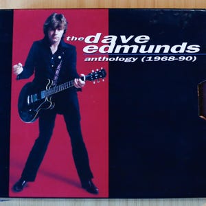 May include: A music CD case featuring a photograph of a musician in a black suit playing a black electric guitar against a red background. The text on the right side reads: the dave edmunds anthology (1968-90) in white and blue lettering.