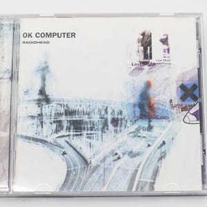 May include: A clear plastic CD jewel case containing the Radiohead album OK Computer. The cover art features a stylized, blurry blue and white illustration of a highway interchange with a dark blue X symbol and the text OK COMPUTER RADIOHEAD.