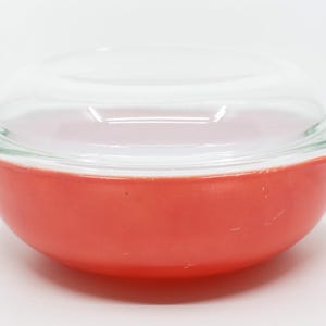 May include: A round, red Pyrex bowl with a clear glass lid. The bowl has a classic design with a slightly flared rim and a smooth, glossy finish. The lid fits snugly on top, creating a complete and functional kitchenware piece.
