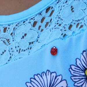 Pictured is a realistic, life-size ladybug pin pinned on a light blue summer dress with lace and daisies modeled by a girl with a medium brown skin tone.