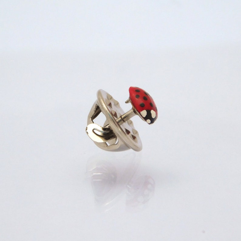 Pictured is a red ladybug pin with a silver-colored back on a reflective light grey background.